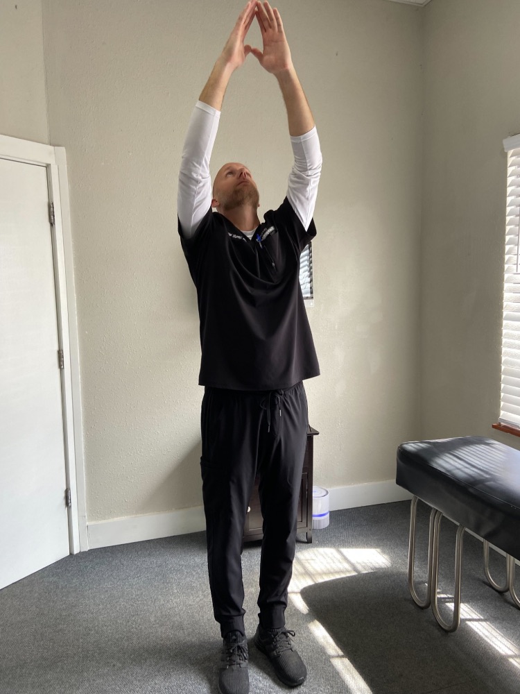 Image of Dr. Daniel Murray: A man is standing up and has his arms up in the air with his hands pressed lightly together.